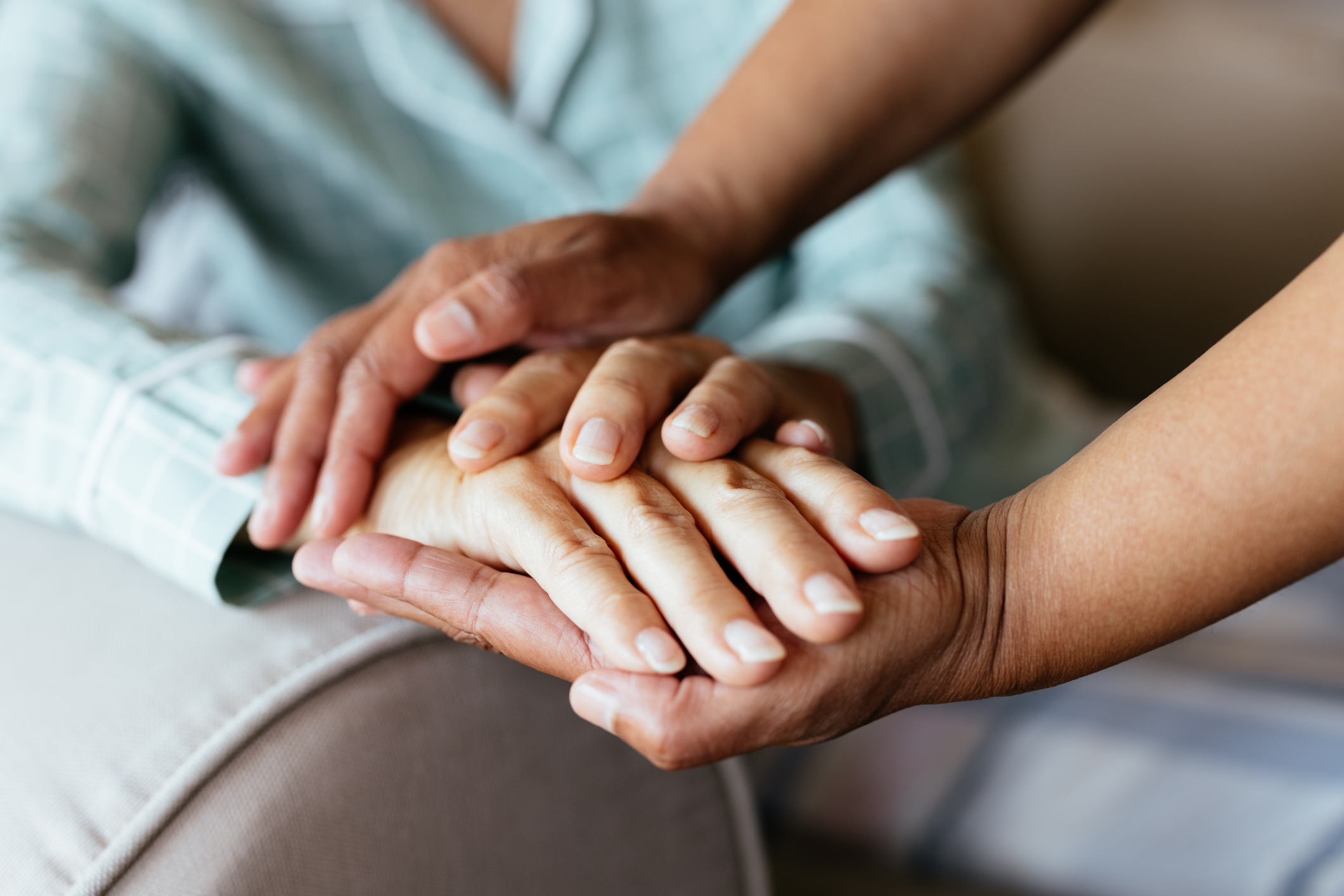 A close-up of a senior woman’s hand being gently held by a nurse or caregiver wearing blue medical scrubs with a stethoscope. The elderly woman wears a green checkered pajama sleeve, symbolizing care, support, and medical assistance in a home or healthcare setting.